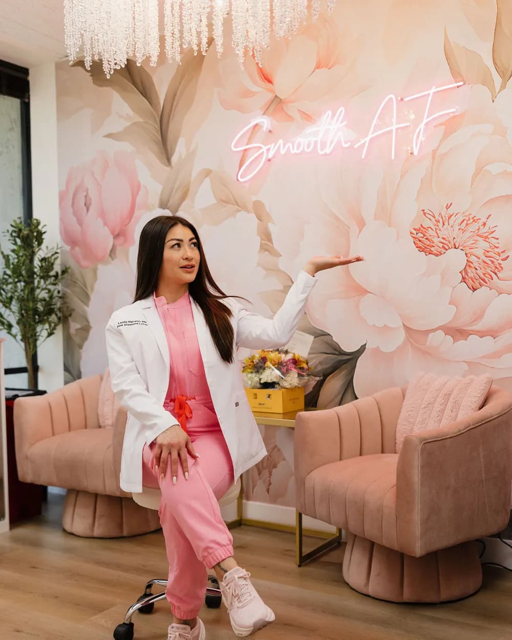 Leslie seated in the Pink Diamond Laser lobby, gesturing at the peony mural under the Smooth AF neon sign